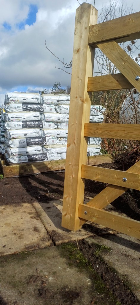 An open wooden gate into the nursery, with stacks of bagged compost on pallets.